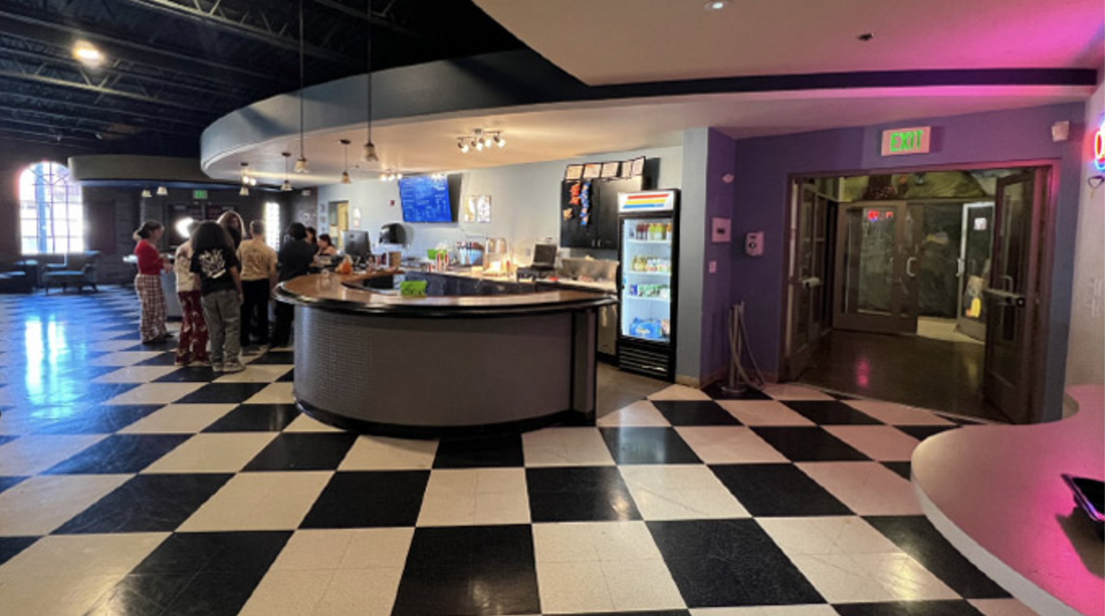 A cafe with a black-and-white checkered floor, round counter, and vending machines. People are lined up at the counter. A doorway is visible on the right.